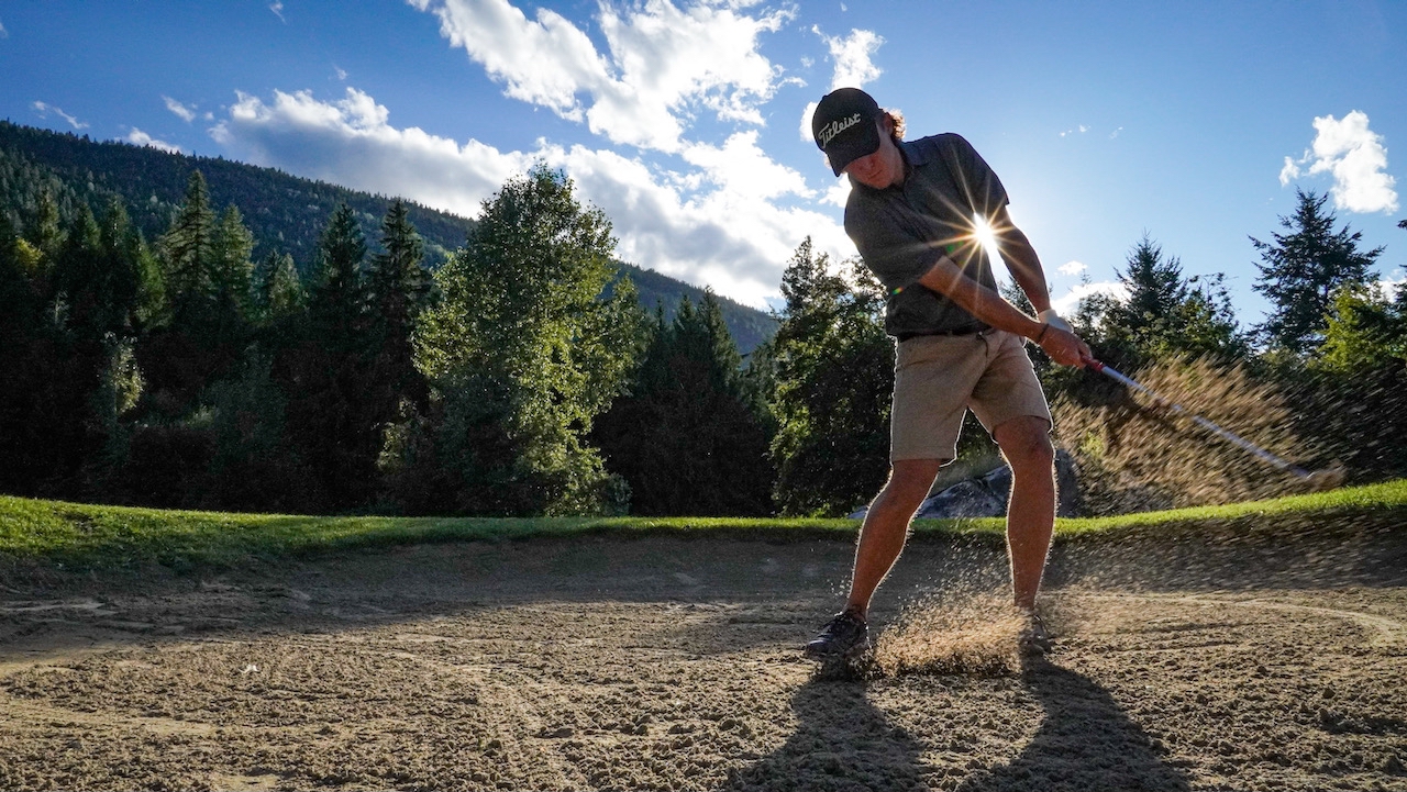 Golfer hitting a ball out of a sand trap at a course in Nelson, BC, with scenic mountains in the background