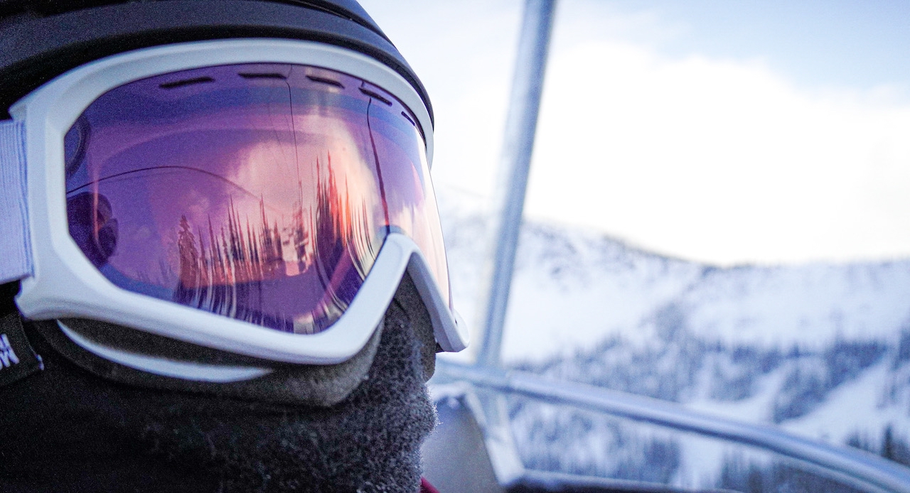 A skier on the lift at Whitewater Ski Resort, wearing a helmet, goggles, and facemask, with snow-covered mountain peaks in the background