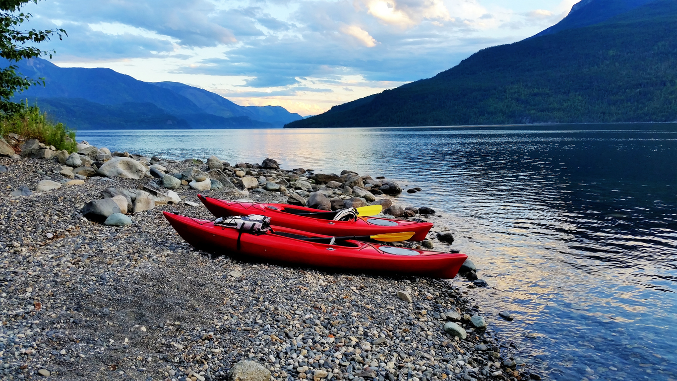 Two kayaks resting on a rocky beach beside Slocan Lake, with calm water and forested mountains in the background.