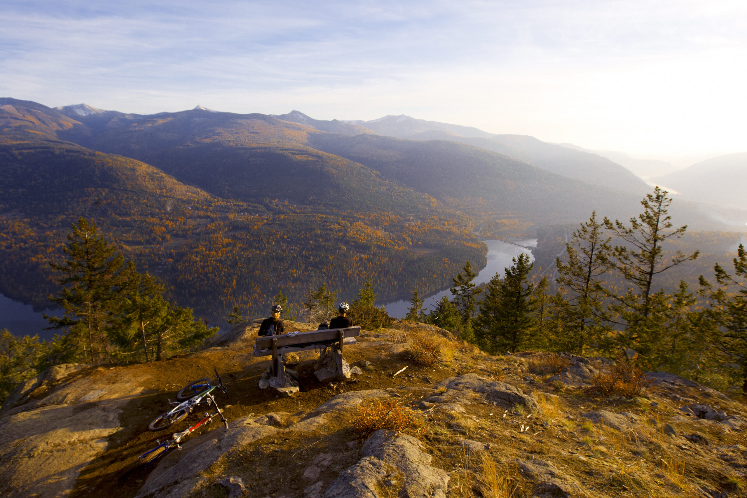 Two mountain bikers relaxing on a bench overlooking Castlegar, with their bikes beside them and the scenic landscape of the Columbia River and surrounding mountains in the background